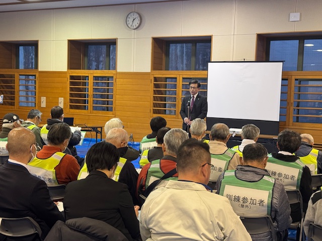 室内で、多くの参加者が「訓練参加者」と書かれた黄緑色のベストを着用して着席しており、前方の男性がマイクを使ってスピーチをしている写真