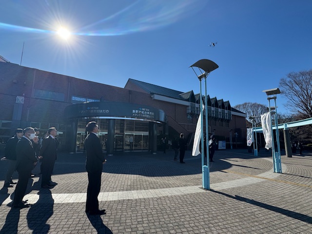 「秦野市総合体育館」の広場で複数のスーツ姿の男性が上空を見上げており、その上空にはドローンらしき飛行物体が浮かんでいる写真