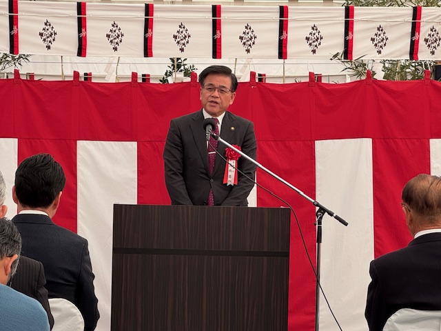 紅白の幕が飾られた会場で、スーツ姿の市長がマイクを使って演台からスピーチをしている様子をアップで写した写真