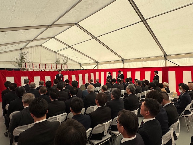 白いテントの中で赤白の幕に囲われた広い会場で、多くのスーツ姿の男性が椅子に座って前方を見ており、その中央の演台に立つ男性がスピーチをしている式典の様子を捉えた写真