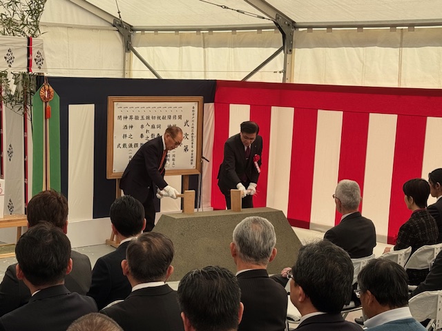 屋外テント内の安全祈願祭で、スーツ姿の男性2名が木槌を手に砂の台を叩いており、その様子を多くの来賓が着席して見守っている写真