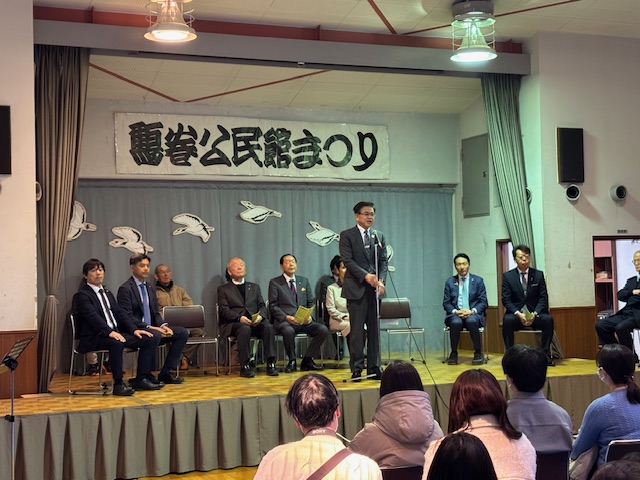 地域のイベントで、ステージ上のマイクを前にスーツ姿の市長がスピーチをしており、多くの聴衆が着席して見ている会場の写真