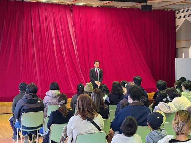 赤い緞帳を背景にしたステージで、スーツ姿の市長がマイクに向かって話しており、多くの人が椅子に座ってそのスピーチを聞いているイベント風景の写真