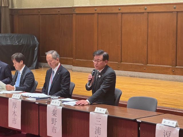 「秦野市」と書かれた名札が置かれたテーブル席で、スーツ姿の市長がマイクを持って発言しており、他の男性たちが隣に座っている会議風景の写真