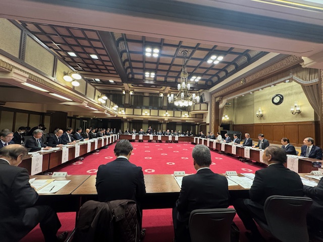 赤い絨毯が敷かれた広い室内の壁際に設置された机に、一人一人名札が貼られ、会議が行われている様子全体を写した写真