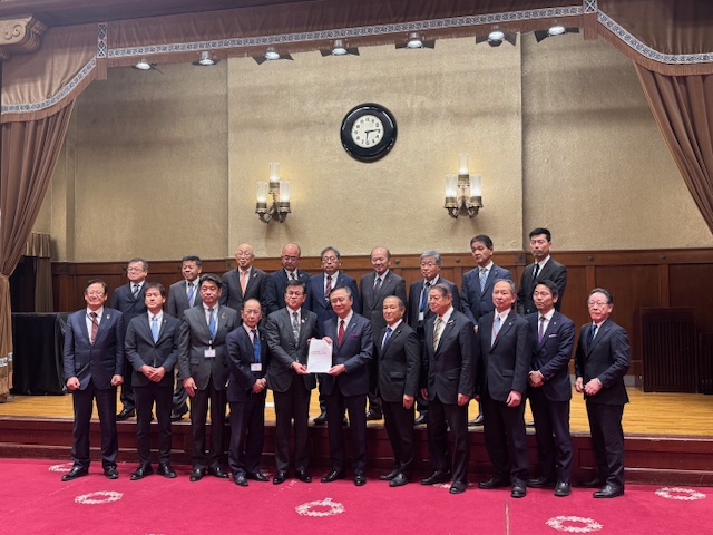 重厚な内装のホールで、スーツ姿の多くの男性たちが中央に集まっており、そのうちの2人が書類を一緒に持ち、カメラに向かって並んでいる集合写真