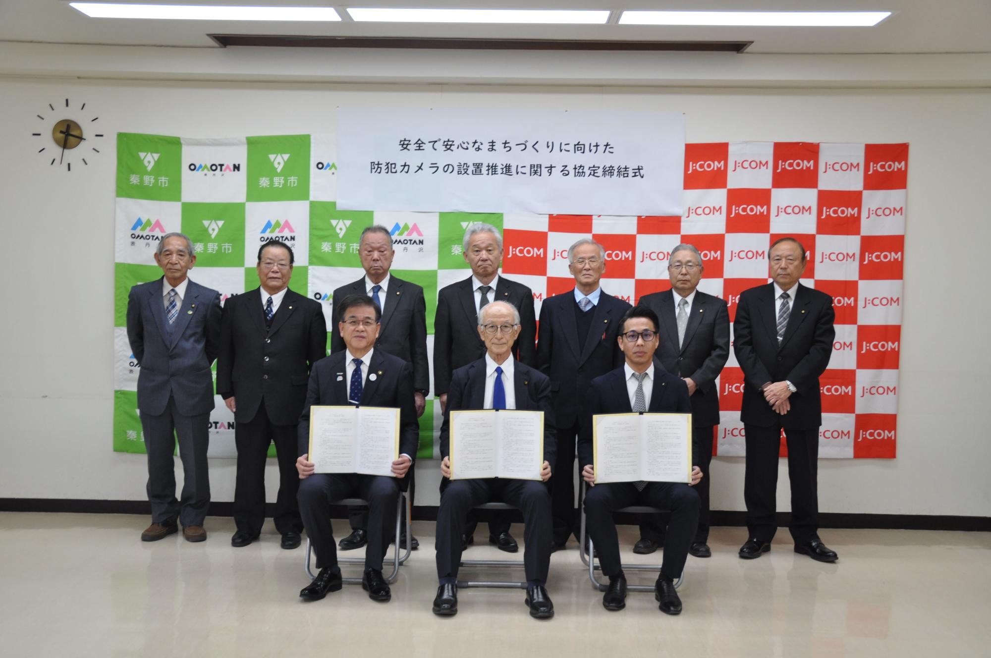 バックパネルの前に立つ10人の男性のうち3人が座って協定書をカメラに向かって掲げている記念写真
