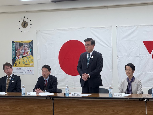 国境が飾られた室内で、スーツ姿の市長がマイクなしでスピーチをしており、その横に男女の関係者がテーブルを囲んで座っている写真