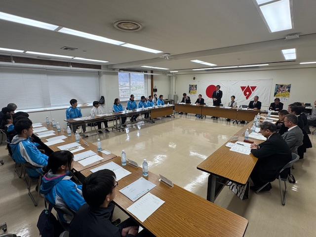 大きなロ字型のテーブルを囲んで複数の選手とスーツ姿の大人たちが座っており、正面の男性がスピーチをしている結団式での写真