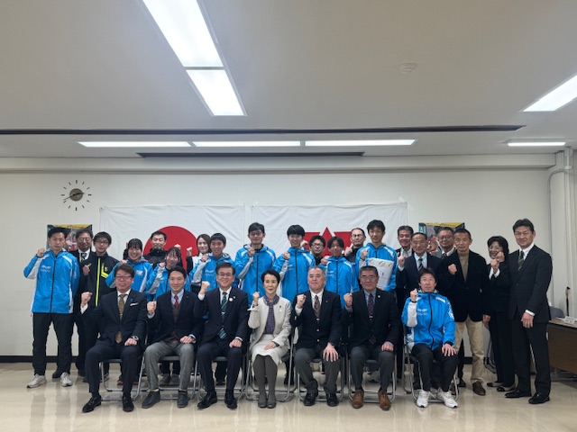 7名が1列目の椅子に座り、他の人たちが後列に並びかながわ駅伝競走大会の選手と関係者の人たちが右手でガッツポーズをして記念撮影をしている結団式での写真