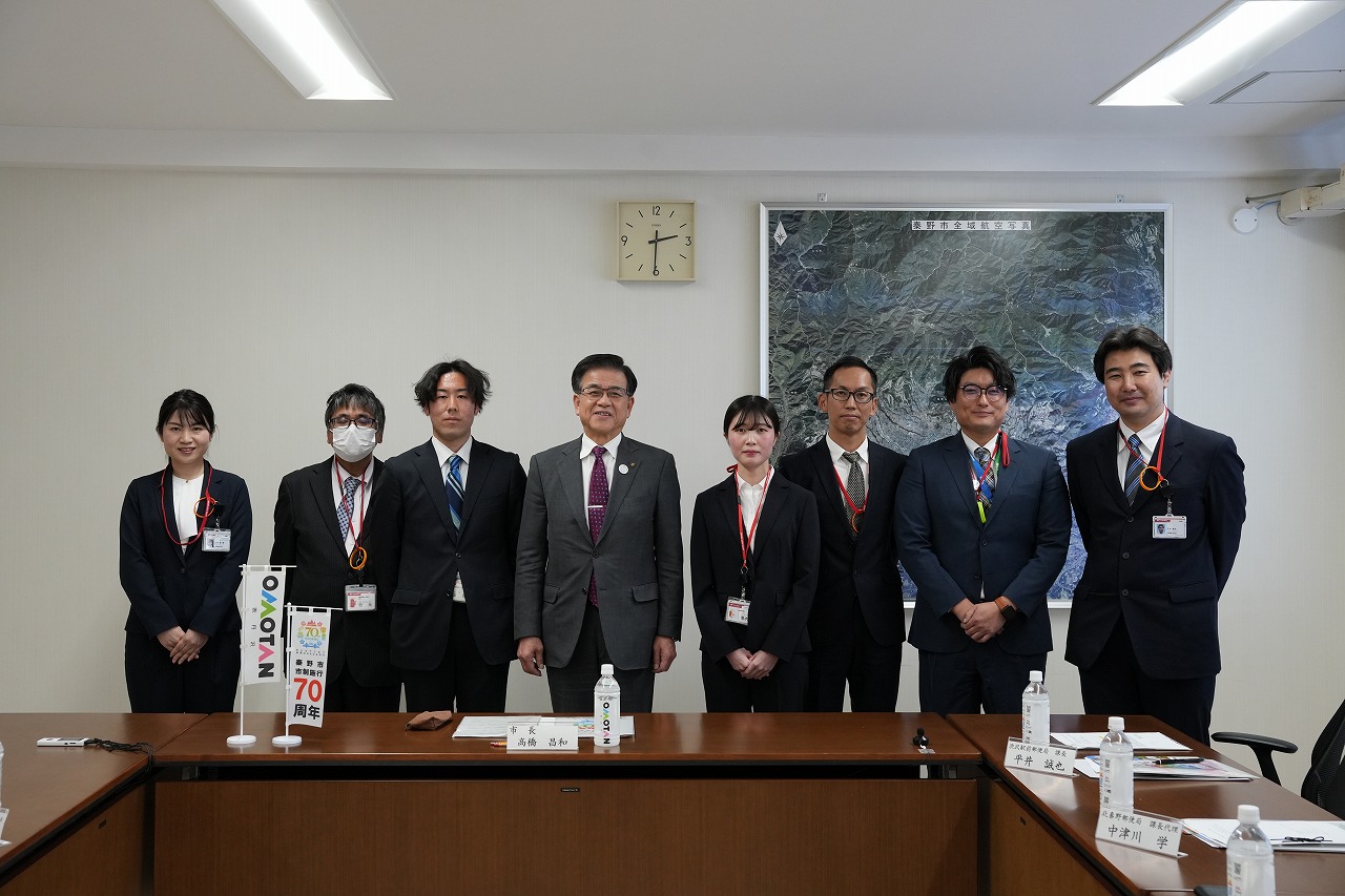 室内で、スーツ姿の男女8人がテーブル名前に並び、カメラの方を向いて記念撮影をしている集合写真