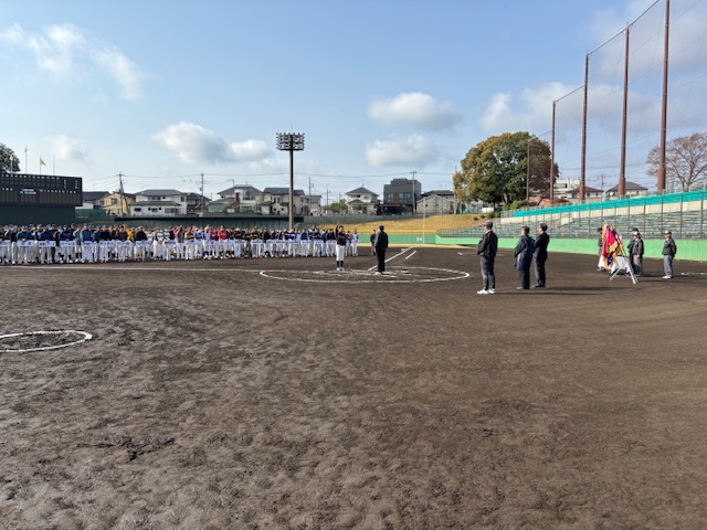 少年野球場のグラウンドで、多くのユニフォーム姿の選手たちが整列し、中央の男性にむかって選手宣誓をしている様子を遠景から撮影した写真