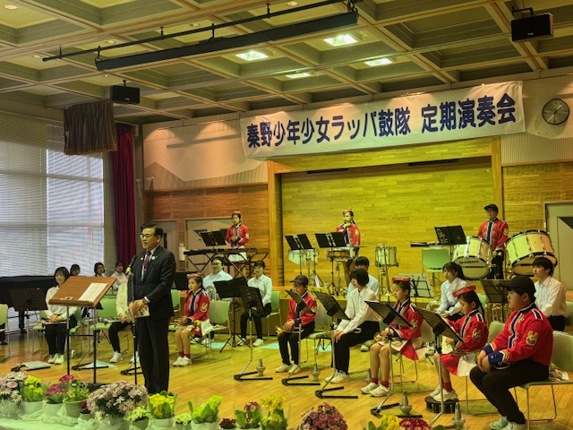 ステージ上で、スーツ姿の男性がスピーチをしており、その周囲には赤い制服や白いシャツに黒いパンツを着た少年少女が楽器の前に座っている演奏会での写真