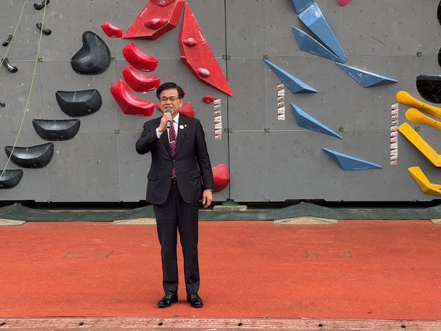 赤い人工芝の上でスーツ姿の男性がマイクを持ってスピーチをしており、その背景には色とりどりのホールドが取り付けられたボルダリングウォールがある写真