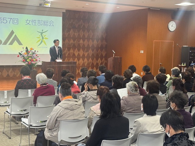 マイクの置かれた演台に立つスーツ姿の男性が、多くの女性の聴衆を前にしてスピーチをしており、背景には「第57回 女性部総会」の文字が映し出されている会場の写真