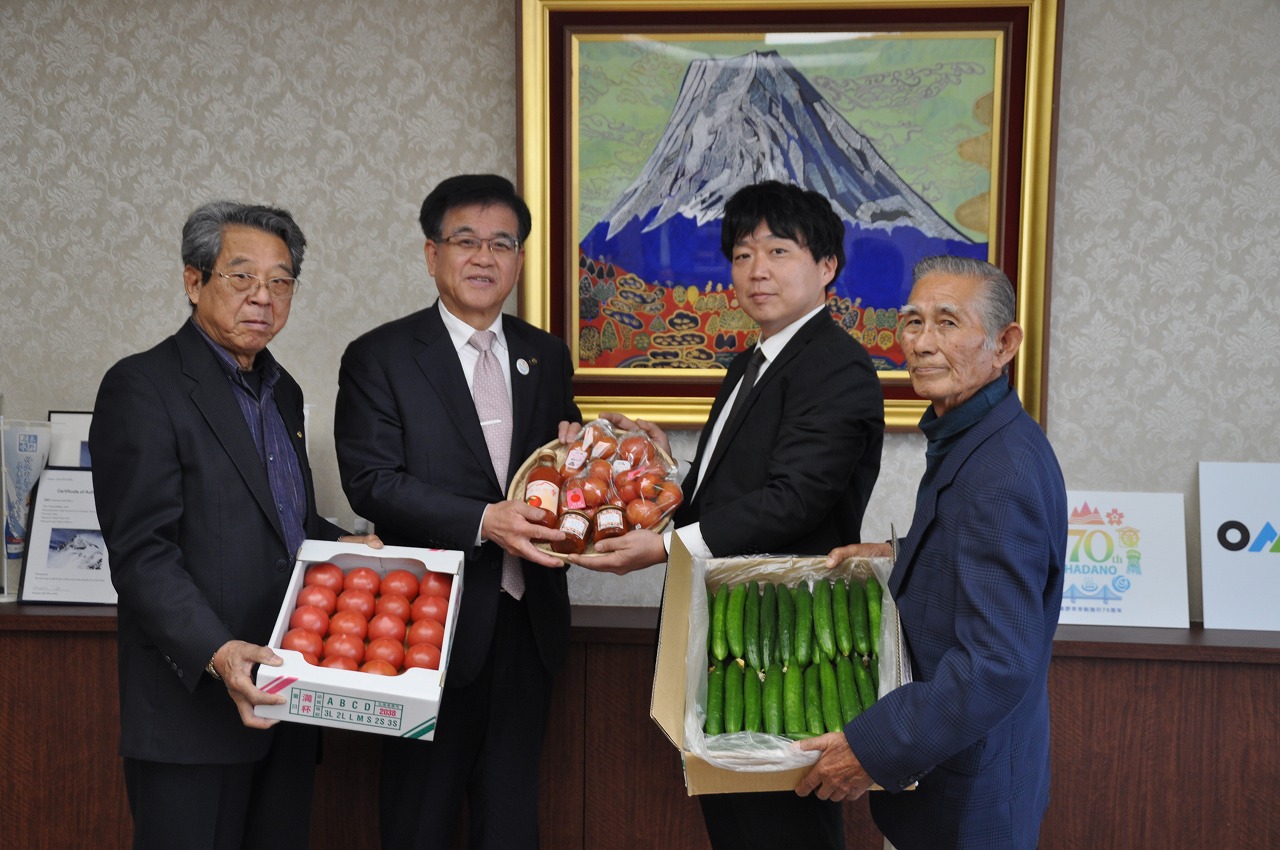 背景に富士山の絵画が飾られているオフィスの応接室で、スーツ姿の4人の男性が、トマトとキュウリの入った段ボール箱や籠を持って笑顔でカメラに並んでいる写真