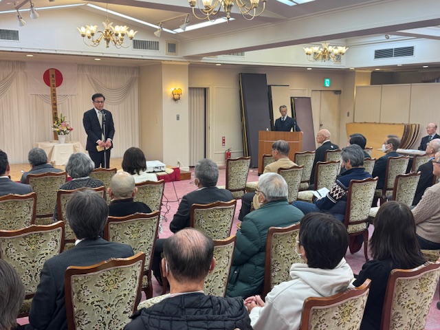 広間のステージ上でスーツ姿の男性がマイクに向かってスピーチをしており、複数の聴衆が椅子に座って話を聞いている様子の写真