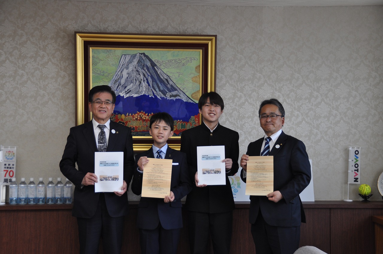 富士山の絵画が飾られた応接室で、スーツ姿の男性2人と学生服の少年2人がそれぞれ賞状を持ってカメラに向かって並んでいる記念写真