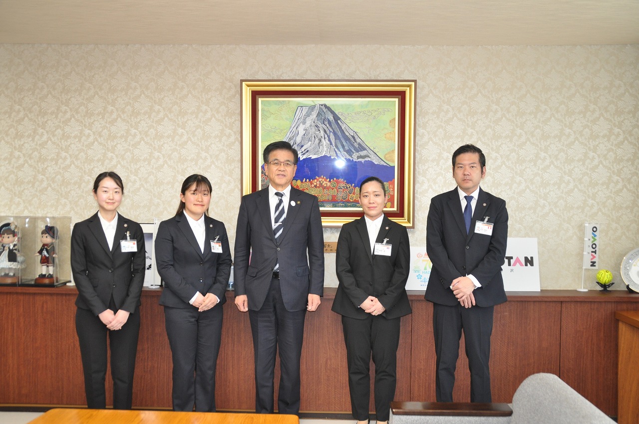 背景に富士山の絵画とキャラクターの置物が飾られている応接室でスーツ姿の男性3人と女性2人が並んでいる集合写真