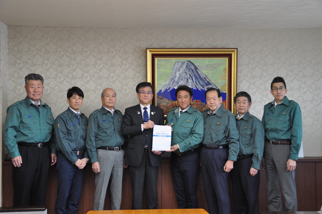 富士山の絵画が飾られた応接室で、スーツ姿の男性を中央に緑色の作業服を着た複数の男性が並んでおり、中央の男性二人が感謝状らしき書類をカメラに向かって掲げている集合写真