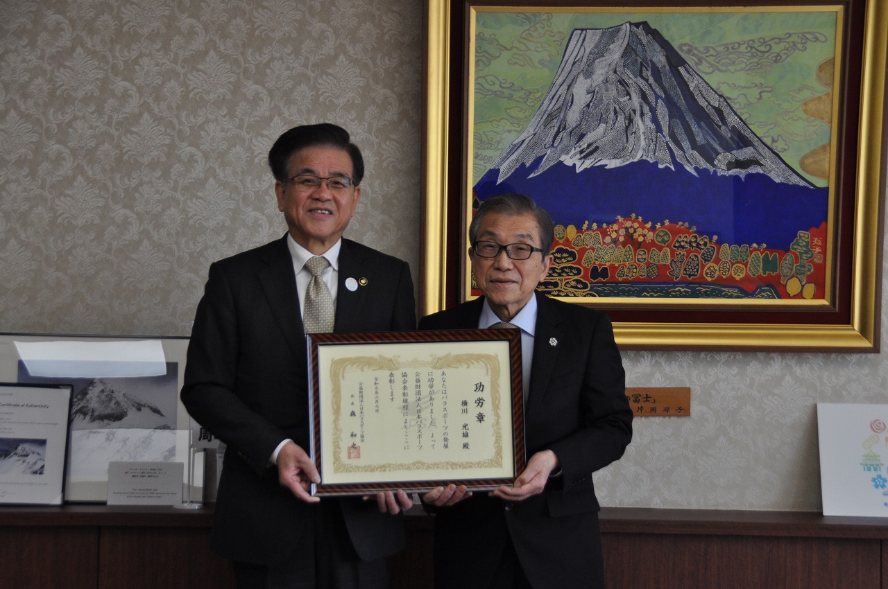 富士山の絵画が飾られた応接室で、スーツ姿の男性二人が勲章と賞状の入った額縁を両手で持ってカメラに向かい、笑顔で並んでいる記念写真