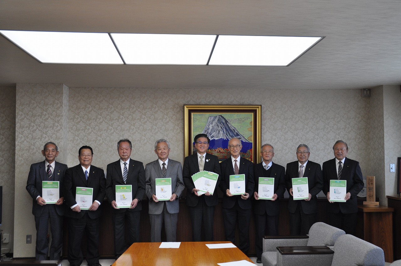 オフィスの応接室で、スーツ姿の9人の男性が冊子や書類を掲げて笑顔でカメラに並んでおり、背景には富士山の絵画が飾られている集合写真