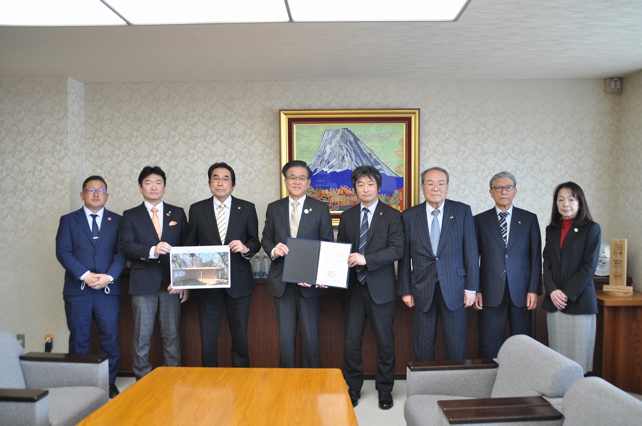 富士山の絵画が飾られた応接室で、スーツ姿の男女8人が並んでおり、中央の男性2人が感謝状と写真を持って笑顔でカメラに並んでいる記念写真