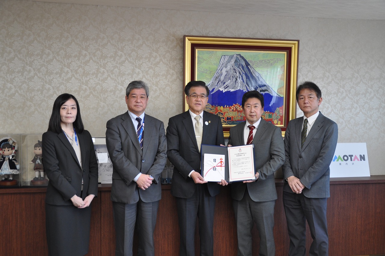 応接室でスーツ姿の男性4人と女性1人が並んでおり、中央の男性2人がお祝いの品と感謝状をカメラに向かって一緒に持っている集合写真