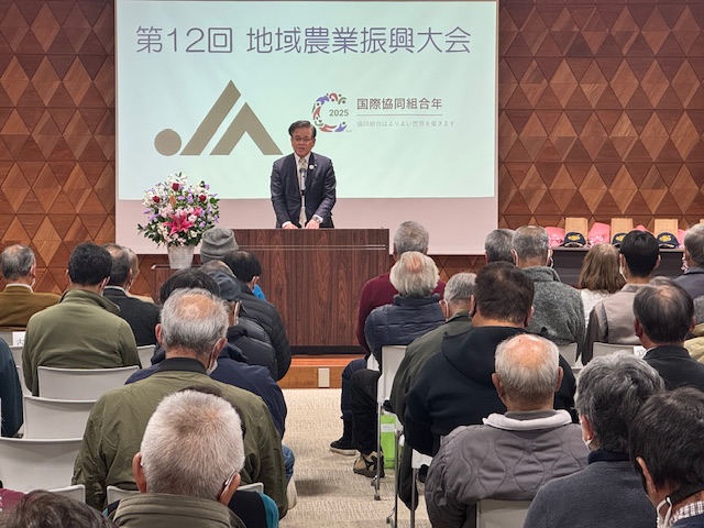 第12回地域農業振興大会の会場で、スーツ姿の男性が聴衆を前に演台から挨拶をしており、背景のスクリーンにはロゴと「国際協同組合年」の文字が映し出されている写真
