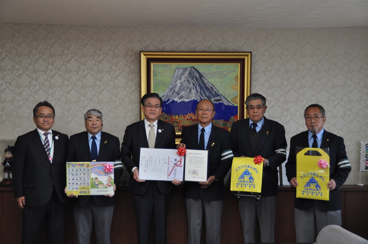 応接室でスーツ姿の6人の男性が並んでおり、中央の3人が賞状と目録を、右端の2人が黄色いバッグをカメラに向かって掲げている集合写真