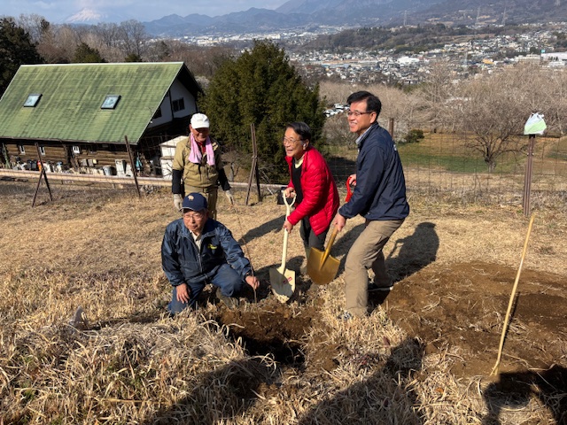 斜面になった屋外の畑で、男女1名ずつがスコップを使い、土をかぶせる作業をし、近くで作業服姿の男性が苗をもち一緒に笑顔を見せている植樹祭での写真