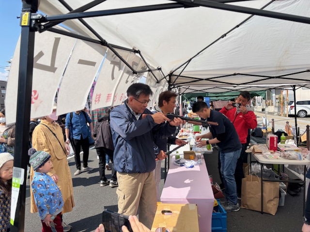 露店の射的場で、男性がライフル銃を構え、その横に立つ別の男性も手伝っており、射的を楽しんでいる様子を撮影した屋外での写真