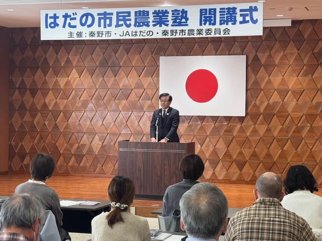 茶色の壁と日の丸が飾られた会場の演台に立つ男性が、着席した多くの聴衆を前にスピーチをしており、上部には「はだの市民農業塾開講式」と書かれた横断幕がある写真