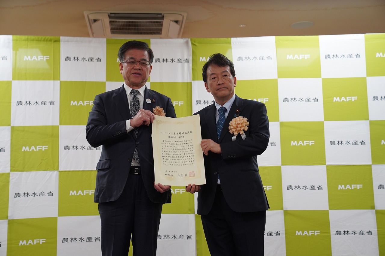 農林水産省と書かれた緑と白の市松模様のパネルを背景に、スーツ姿の男性二人が感謝状を一緒に持ち、笑顔でカメラに向かっている記念写真