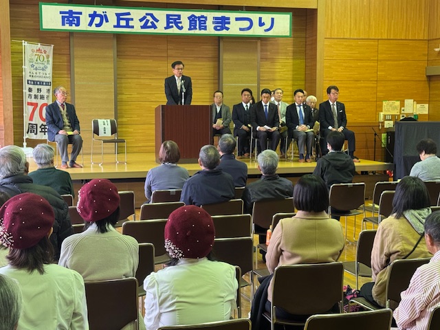 南が丘公民館まつりの横断幕が飾られ関係者が座っている舞台の演台でスーツ姿の男性が挨拶をしている様子を写した写真