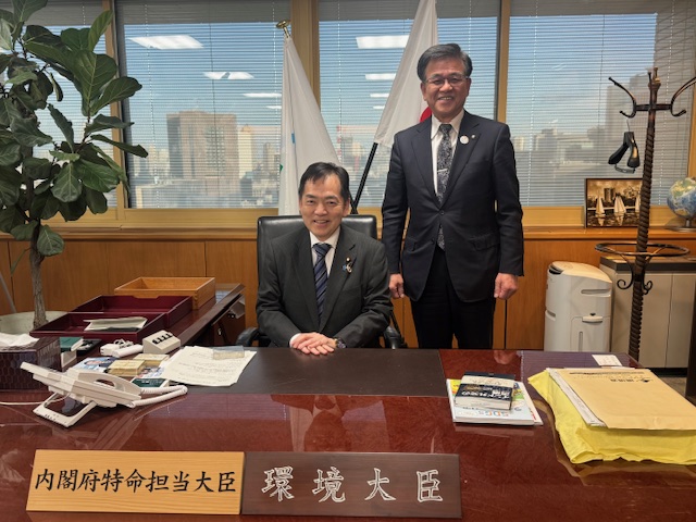 机に座っている浅尾慶一郎環境大臣と一緒に記念撮影をしている市長の写真