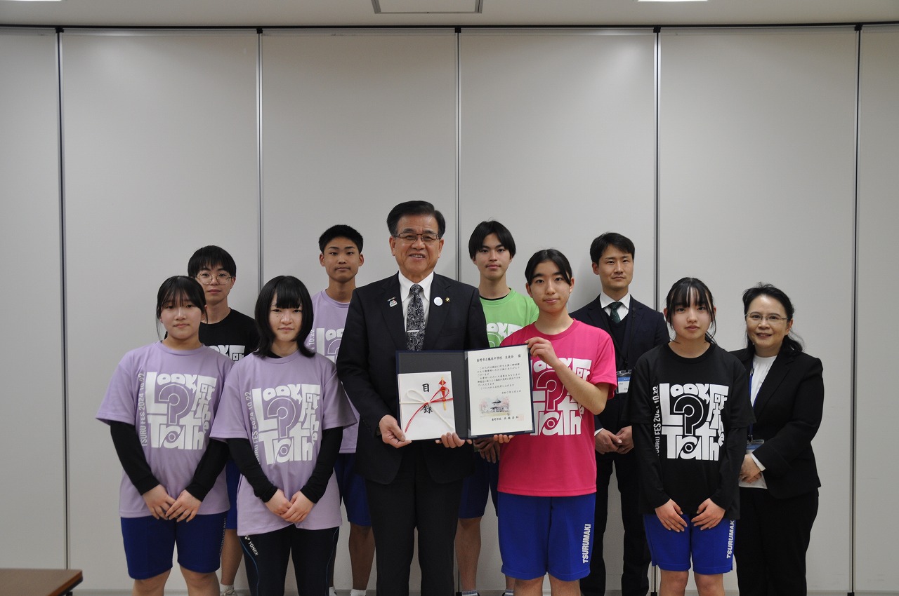 スーツ姿の男女と部活動のユニフォームを着た少年少女が、感謝状や目録を囲んで笑顔でカメラに並んでいる集合写真