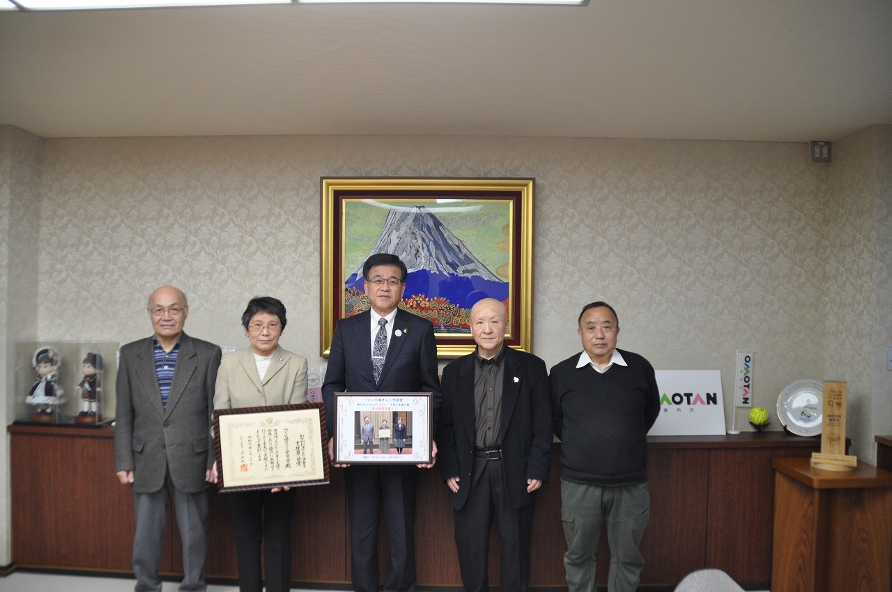 市役所の応接室で、スーツ姿の男性を中央に5人が並んでおり、男性と女性が感謝状と写真の入った額縁をカメラに向かって一緒に持っている記念写真