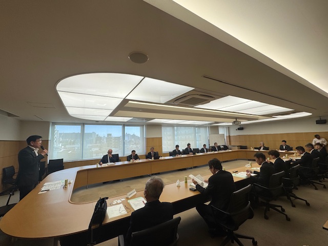 複数の男性がロの字型のテーブルを囲んで座る会議室で、市長がマイクを持ちながら発言している様子の写真