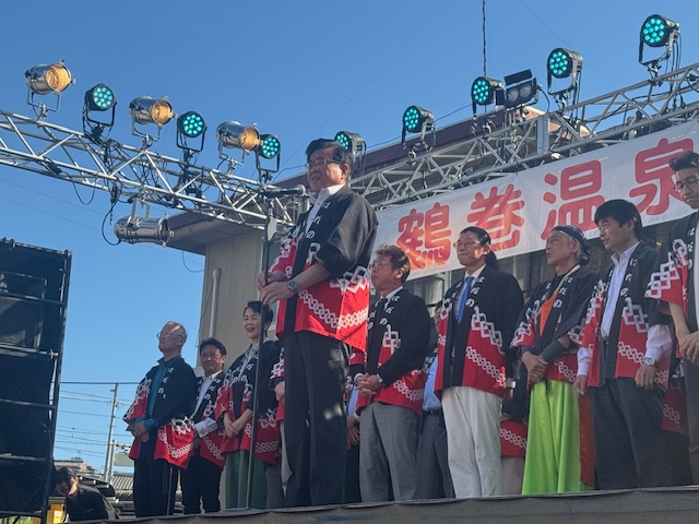 ステージに上がり「鶴巻温泉」と書かれた看板を背景に、お揃いの法被を着た人々に囲まれながらマイクで挨拶をする市長の写真