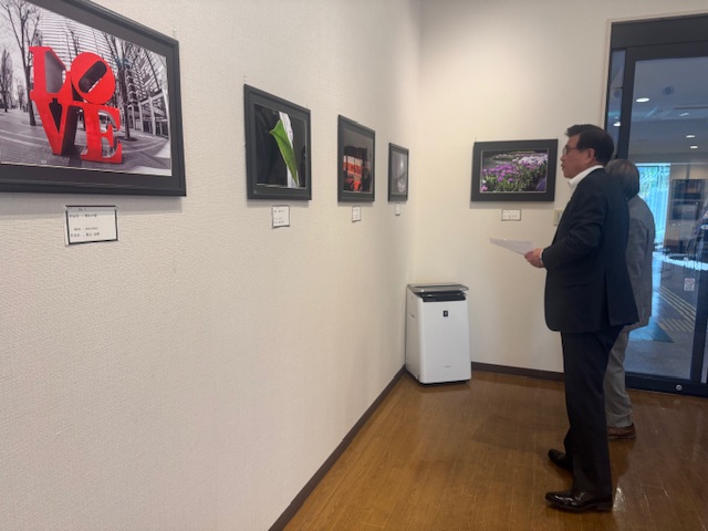 展示スペースの白い壁に飾られた複数の写真作品を、黒いスーツ姿の市長が鑑賞している様子の写真