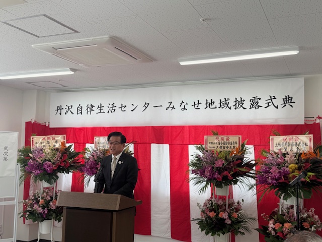 「丹沢自立生活センターみなせ地域披露式典」と書かれた垂れ幕の前で、演台に立ち花輪に囲まれながら挨拶する黒いスーツ姿の市長の写真