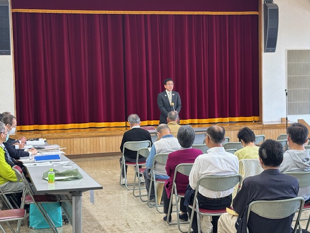 講堂のようなステージの前でマイクを持ち、椅子に座った聴衆に向かって話をしているスーツ姿の市長の写真