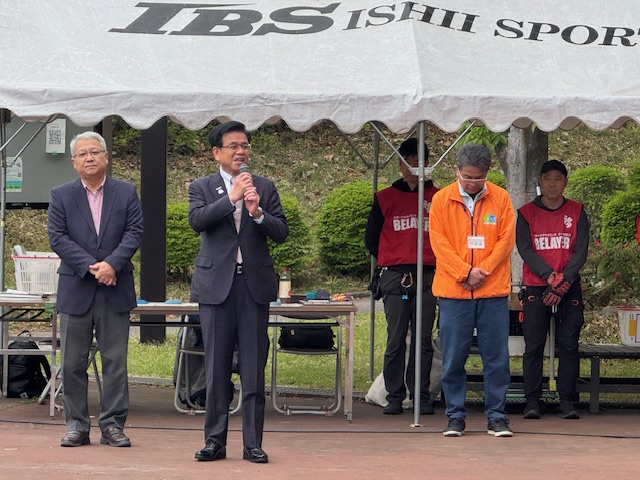 黒いスーツ姿の市長がマイクを手にスピーチをしており、その両脇には別の男性が立ち、背景には「IBS ISHII SPORT」のロゴが入った白いテントとスタッフの姿が見える式典の様子を捉えた写真