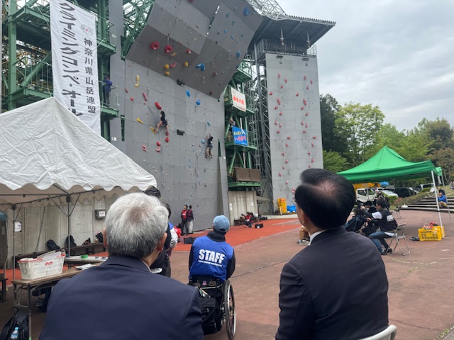 カラフルなホールドが設置されたそびえ立つクライミングウォールで、競技者が挑戦している様子を、多くの人々が座ったり立ったりしながら見守り、ボランティアスタッフもいる、屋外でのイベントの様子を捉えた写真