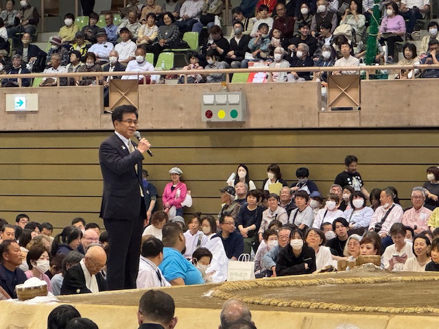 黒いスーツを着た市長がマイクを片手に土俵のそばで観客席に向かってスピーチをしており、その周囲には多くの老若男女の観客が座って見ている、相撲会場でのイベントの様子を捉えた写真