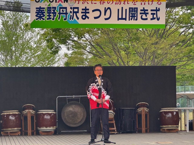 丹沢まつり 山開き式の横断幕の下で法被姿の市長がマイクを使い挨拶をしている写真