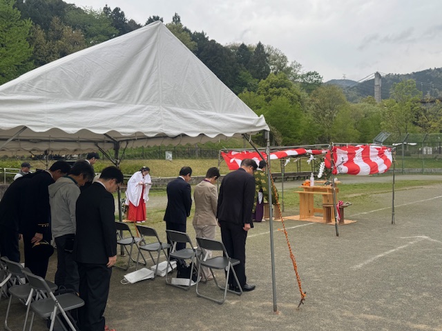 広い敷地のテントの下で、スーツ姿の男性たちや巫女が神事のために一列に並んで深くお辞儀をしており、手前には祭壇のようなものが設置されている、行事の様子を捉えた写真