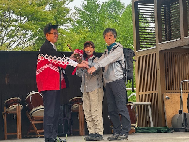 赤い法被を着た市長が、登山服を着た若い男女に大きな鍵のようなものを手渡している山開き式での写真