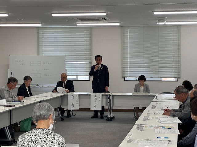 多くの人々が机に向かい、スーツ姿の男性がマイクを手に立って話をしており、その周囲には複数の男女が着席し、ホワイトボードや名前が書かれた札が見える会議の様子を捉えた写真